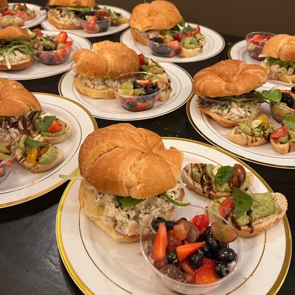 Plates of chicken salad croissant sandwiches, fruit cups, and avocado bruschetta on a dark table.