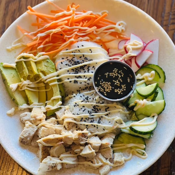A colorful poke bowl with chicken, avocado, carrots, cucumbers, radishes, rice, and sauce.