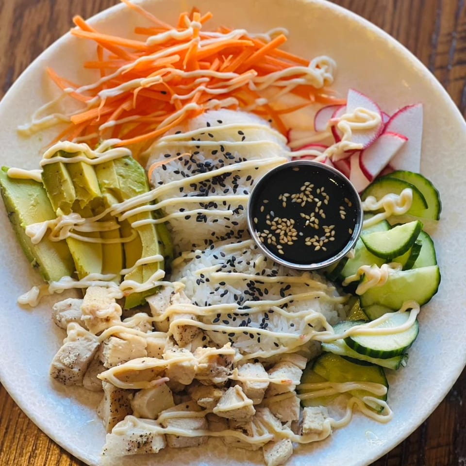 A colorful poke bowl with chicken, avocado, carrots, cucumbers, radishes, rice, and sauce.