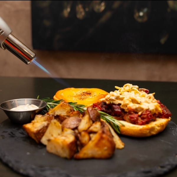 A kitchen torch caramelizing a dish with roasted potatoes, meat, and cheese on a dark slate.