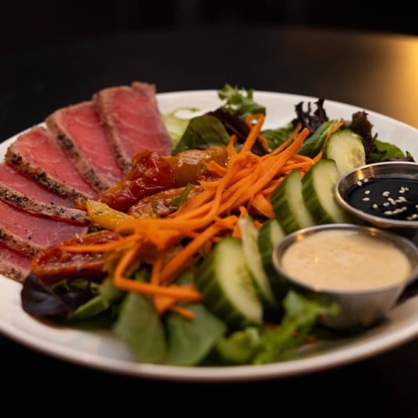 Seared tuna slices with mixed greens, shredded carrots, cucumbers, and two dipping sauces on a white plate.
