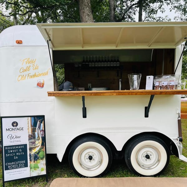 Montage Wine Bar's white mobile wine tasting trailer with a wooden counter and bottles inside.