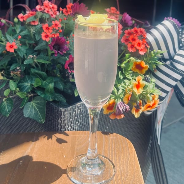 A champagne flute with a light pink drink and lemon garnish sits on a wooden table amidst colorful flowers.