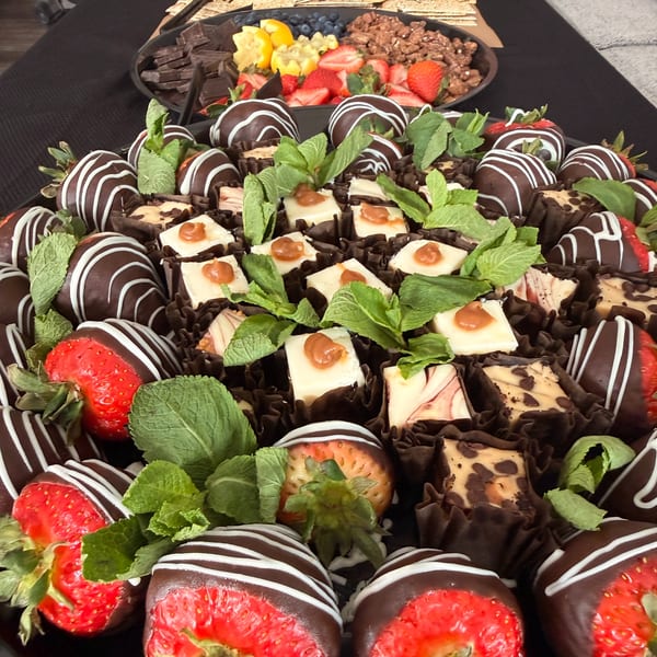 A platter of chocolate-covered strawberries, cheesecake bites, and mint garnishes, with a fruit and chocolate tray in the background.