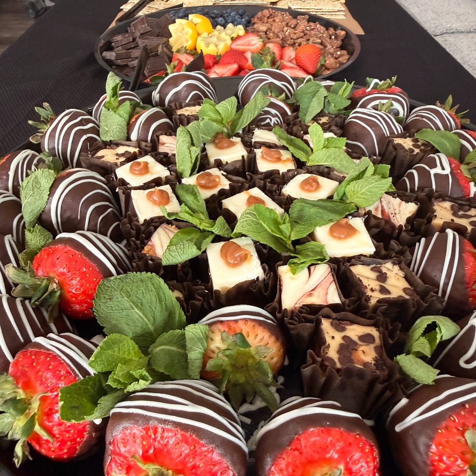 A platter of chocolate-covered strawberries, cheesecake bites, and mint garnishes, with a fruit and chocolate tray in the background.