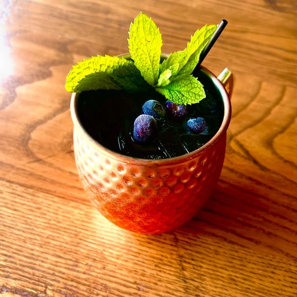 A copper mug cocktail with blueberries, mint, and a black straw on a wooden table.
