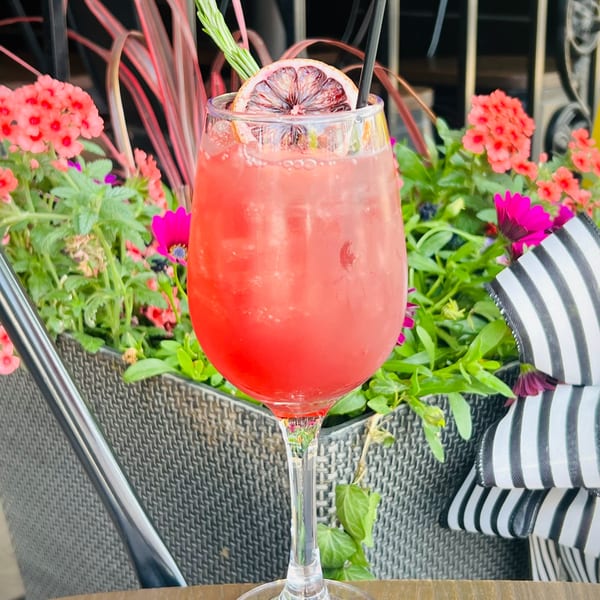 Pink cocktail with a blood orange slice and rosemary garnish, surrounded by vibrant flowers.