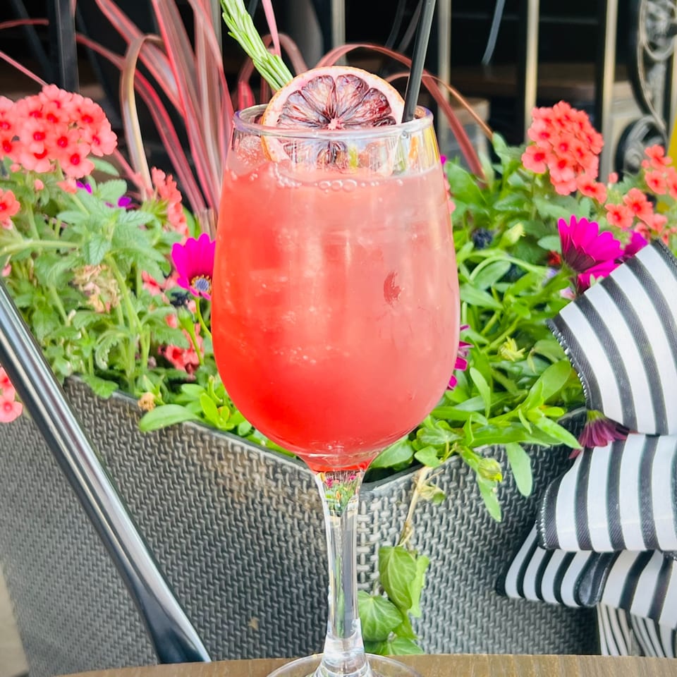 Pink cocktail with a blood orange slice and rosemary garnish, surrounded by vibrant flowers.